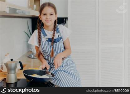 Indoor shot of pleasant looking seven years old girl busy frying egss, holds pan and sits near stove, dressed in apron, looks positively at camera, boasts she can cook, poses in modern kitchen