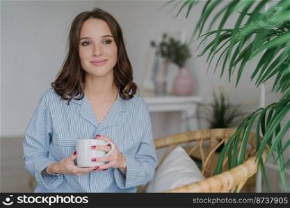 Indoor shot of pleased attractive woman with dark hair, healthy soft skin, drinks coffee in morning, wears pyjamas, looks thoughtfully aside, poses at cozy room, feels relaxed. Leisure concept