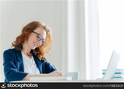 Indoor shot of pleased cheerful famale with ginger hair, signs contract, sits at cabinet in front of laptop computer. Smiling director notes information for financial report, works on laptop computer