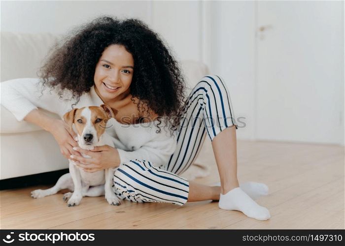 Indoor shot of pleased curly haired female model poses for photo with jack russell terrier dog, embraces pet tenderly, sits on floor with beloved animal near comfortable sofa, spend time together