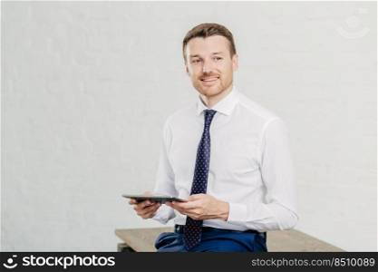 Indoor shot of pleased male owner of prosperous company dressed in formal clothes, holds modern tablet computer, enjoys online communication, looks joyfully aside, has bristle, poses in office