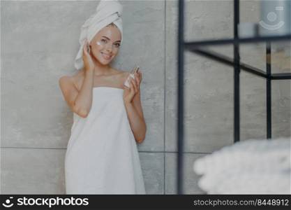 Indoor shot of pleased young European woman applies cosmetic product on face wrapped in bath towel has healthy glowing skin poses against grey background at bathroom. Facial treatment concept