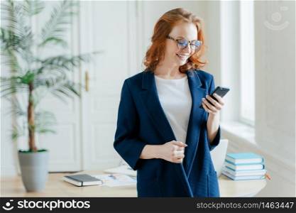 Indoor shot of positive red haired woman checks emial box on modern smartphone device, wears optical glasses, uses modern technologies, recieves good news, chats online, dressed in formal wear
