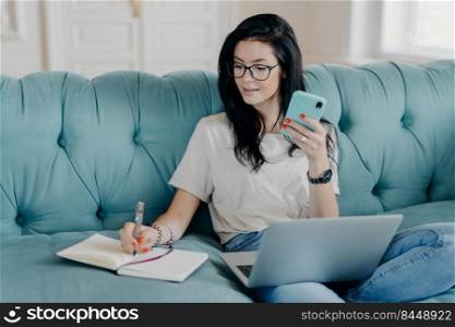 Indoor shot of pretty brunette female student watches webinar and makes notes in notepad, holds mobile phone, sits on comfortable sofa, busy with distance job. People, technology, lifestyle concept
