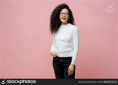 Indoor shot of pretty cheerful woman going to work, laughs at something positive, wears neat white jumper and jeans, keeps hand in pocket, has fun with friends during free time, poses indoor