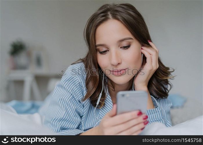 Indoor shot of pretty dark haired woman enjoys music in playlist, lies on comfortabled bed at home, has day off, watches films online, connected to wireless internet, enjoys cozy atmosphere.