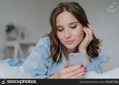 Indoor shot of pretty dark haired woman enjoys music in playlist, lies on comfortabled bed at home, has day off, watches films online, connected to wireless internet, enjoys cozy atmosphere.