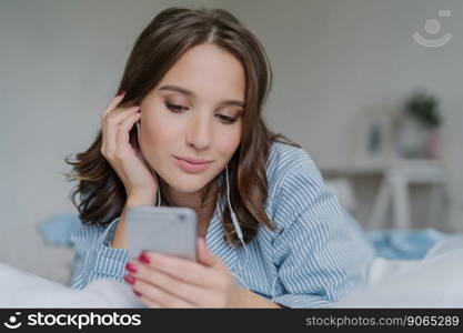 Indoor shot of pretty dark haired woman enjoys music in playlist, lies on comfortabled bed at home, has day off, watches films online, connected to wireless internet, enjoys cozy atmosphere.