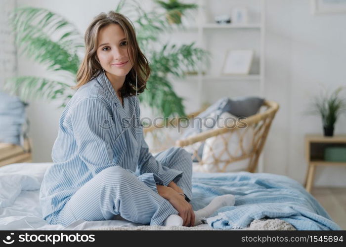 Indoor shot of pretty dark haired woman sits crossed legs on bed, dressed in nighclothes, white socks, looks direcltly at camera, feels relaxed while listens calm music in earphones. Rest and day off