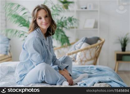 Indoor shot of pretty dark haired woman sits crossed legs on bed, dressed in nighclothes, white socks, looks direcltly at camera, feels relaxed while listens calm music in earphones. Rest and day off