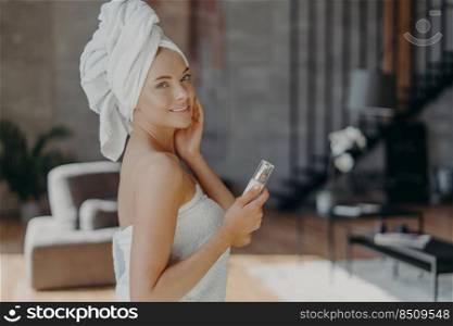 Indoor shot of pretty European woman with healthy glowing skin, minimal makeup, holds bottle of body lotion, p&ers complexion, wears bath towel around body, poses against domestic interior