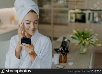 Indoor shot of pretty young woman applies face cream smiles gently looks at mirror enjoys softness of skin after taking shower wears bathrobe and wrapped towel on head poses in modern apartment