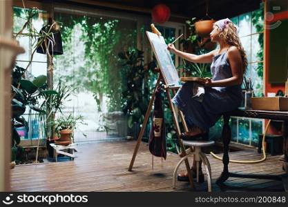 Indoor shot of professional female artist painting on canvas in studio with plants.