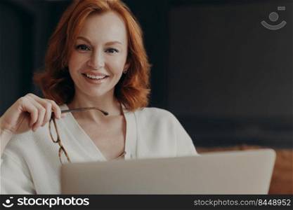 Indoor shot of professional woman copywriter works on creation advertising content, browses internet, smiles gladfully and holds transparent glasses, uses modern laptop computer, works remotely