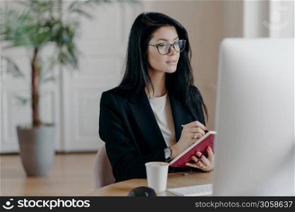 Indoor shot of prosperous businesswoman makes notes in notepad, concentrated in computer screen, watches training video how to start own business, poses at desktop with coffee cup, makes report