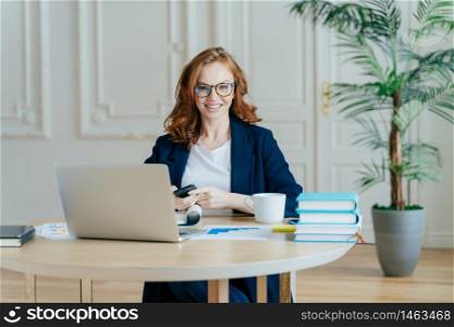 Indoor shot of red haired female economist sends text message to colleague, holds contemporary smart phone, poses at workplace, works with financial documents, surrounded with modern technologies