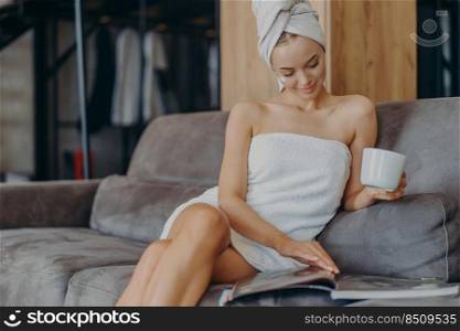 Indoor shot of relaxed beautiful woman sits relaxed on sofa, wears bath towel around body after taking shower, holds cup of tea, reads magazine, spends free time at home. Wellness, hygiene concept