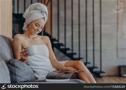 Indoor shot of relaxed beautiful woman wrapped in towel, drinks aromatic coffee from cup, sits on comfortable sofa in living room, reads magazine, spends free time at home. Domestic atmosphere