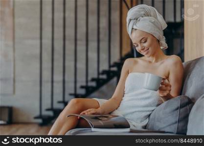Indoor shot of relaxed beautiful woman wrapped in towel, drinks aromatic coffee from cup, sits on comfortable sofa in living room, reads magazine, spends free time at home. Domestic atmosphere