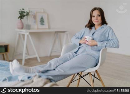 Indoor shot of relaxed female dressed in pyjamas, sits on chair, keeps legs on bed, drinks hot coffee, poses against domestic interior, has appealing look. People, rest and relaxation concept