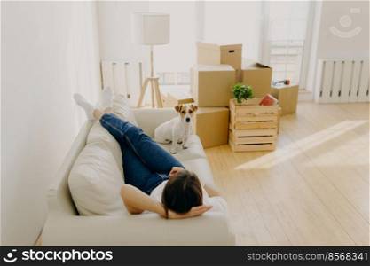 Indoor shot of relaxed woman rests on sofa with favourite pet, removes in new rented apartment, pile of carton boxes and l&near. Female homeowner feels happy of being in new home. Relocation
