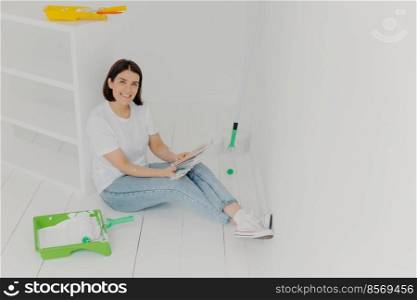 Indoor shot of satisfied brunette housewife sits on floor, studies color s&les, decides best tone for new apartment, wears white t shirt, jeans and sneakers, happy almost to finish house repair