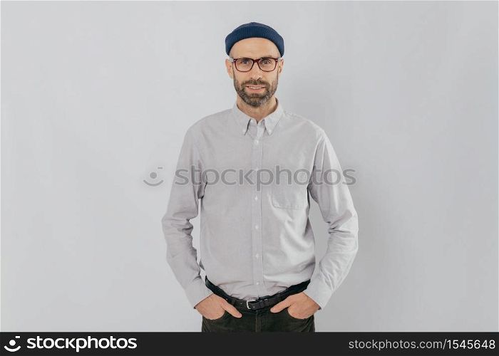 Indoor shot of satisfied Caucasian man in spectacles with bristle, keeps hands in pockets, dressed in formal stylish clothes, has gentle smile, happy to be promoted, isolated over white background.