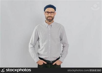 Indoor shot of satisfied Caucasian man in spectacles with bristle, keeps hands in pockets, dressed in formal stylish clothes, has gentle smile, happy to be promoted, isolated over white background.