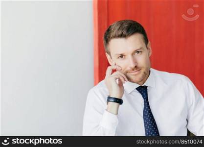 Indoor shot of self confident businessman in white shirt with tie, keeps hands near face, waits for someone, isolated over red and white background. Successful male entrepreneur poses indoor