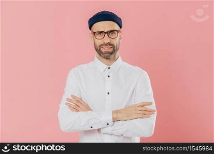 Indoor shot of self confident unshaven man looks happily at camera, keeps arms folded, wears headgear and shirt, listens attentively interlocutor, isolated over pink background. Masculinity concept