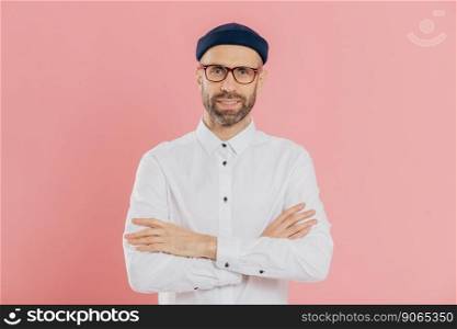 Indoor shot of self confident unshaven man looks happily at camera, keeps arms folded, wears headgear and shirt, listens attentively interlocutor, isolated over pink background. Masculinity concept