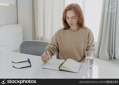 Indoor shot of serious redhead young woman makes notes in notepad makes list to do plans workday manager time sits at white desktop with glass of water spectacles against cozy domestic interior. serious redhead young woman makes notes in notepad sits at table