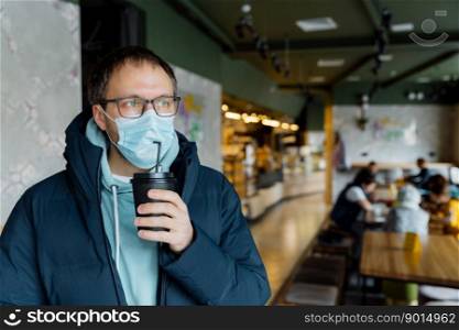Indoor shot of serious thoughtful man drinks takeaway coffee, wears protective mask, transparent glasses, jacket, poses in crowded cafe, follows quarantine rules during coronavirus outbreak.