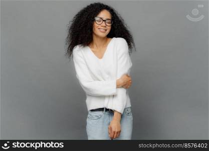 Indoor shot of sincere woman keeps hand partly crossed, dressed in white jumper and jeans, has eyes closed, enjoys happy moment in life, isolated over grey background. Positive emotions concept
