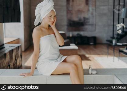 Indoor shot of slim attractive young European woman applies moisturising facial cream, poses in modern apartment, wears towel on head, tries new cosmetic product. Skin care and beauty concept