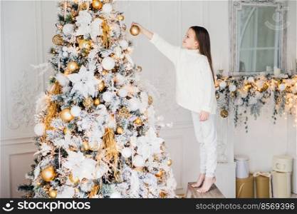 Indoor shot of small kid in white clothes, decorates New Year tree, stands on tiptoe, holds decoration in hands, does everything without parent`s help. House decorating and childhood concept.