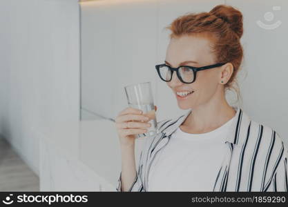 Indoor shot of smiling redhead young woman holds glass of water drinks aqua for body refreshment follows healthy lifestyle wears spectacles striped shirt focused into distance. Hydration concept. Smiling redhead young woman holds glass of water drinks aqua for body refreshment