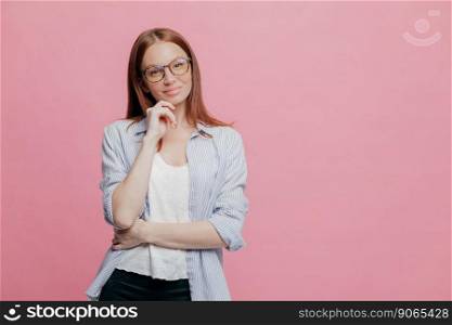 Indoor shot of thoughtful Caucasian woman keeps one hand under chin, dressed in elegant shirt, models over pink background, isolated over pink background with copy space for your promotion or slogan