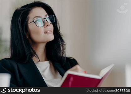 Indoor shot of thoughtful dreamy brunette female jouranlist works on book review, holds opened notepad, writes down some ideas or memo notes, creats new article, works on project, wears glasses.