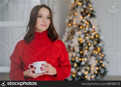 Indoor shot of thoughtful young female in warm winter clothes, enjoys hot coffee or cappuccino, looks pensively aside, stands near decorated Christmas tree, has red manicure. Coziness concept