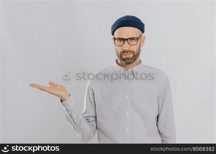 Indoor shot of unshaven Caucasian man holds copy space, demonstrates something against white wall, involved in advertising c&aign. Serious male model with bristle stands alone. Isolated shot