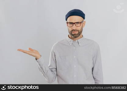 Indoor shot of unshaven Caucasian man holds copy space, demonstrates something against white wall, involved in advertising c&aign. Serious male model with bristle stands alone. Isolated shot