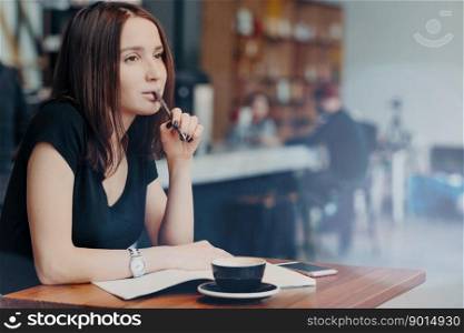 Indoor shot of young female student writes down notes for report, works in coffee shop, drinks cappuccino, plans organization process, has thoughtful expression, creats essay or prepares for exam