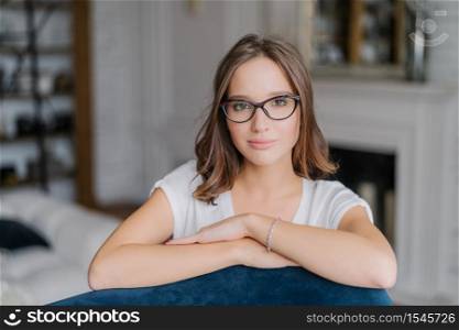 Indoor view of pretty woman in spectacles, keeps hands on back of sofa, poses in living room at home, thinks about something, dressed in casual clothes. People, free time and lifestyle concept