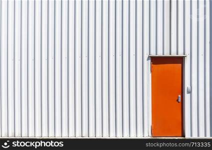 Industrial building facade with a metal orange door and a white aluminum sheet wall. White warehouse with corrugated metal wall and orange door.