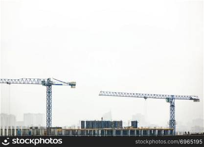 Industrial landscape with silhouettes of cranes on the gray cloudy background. Photo from the drone.. Building under construction with two tower cranes against a gray cloudy sky.