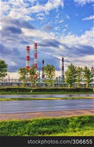 Industrial summer landscape - view across the road and green trees to an oil refinery under a blue dramatic sky with clouds at sunny day.. Oil Refinery At Sunny Day