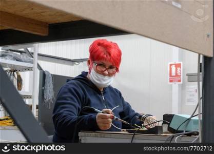 Industrial worker wearing a face mask due to a coronavirus pandemic solders cables from factory production equipment. High quality photo. Industrial worker wearing a face mask due to a coronavirus pandemic solders cables from factory production equipment