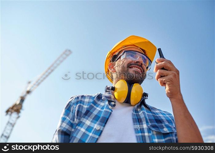 industry, building, technology and people concept - male builder in hardhat with walkie talkie or radio outdoors