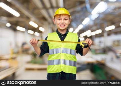 industry, manufacture and profession concept - happy smiling little boy in protective helmet and safety vest with ruler over workshop background. boy in construction helmet and vest with ruler